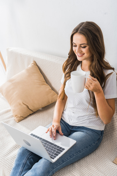 high angle view of female freelancer with coffee cup working with laptop on couch at home  - Foto, Imagen