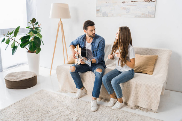 young handsome man playing on acoustic guitar while his girlfriend dancing near on couch at home - 写真・画像