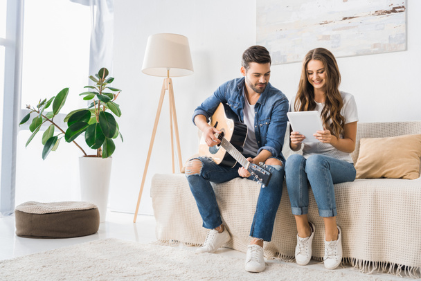 smiling couple tuning acoustic guitar with digital tablet on sofa at home - Photo, Image
