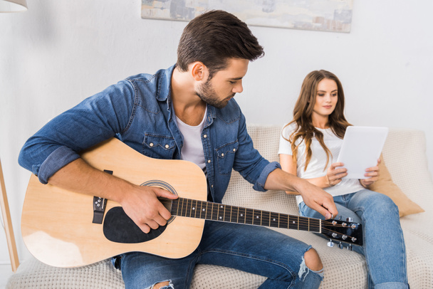 handsome man tuning guitar while his girlfriend sitting near with digital tablet on sofa at home - Photo, Image