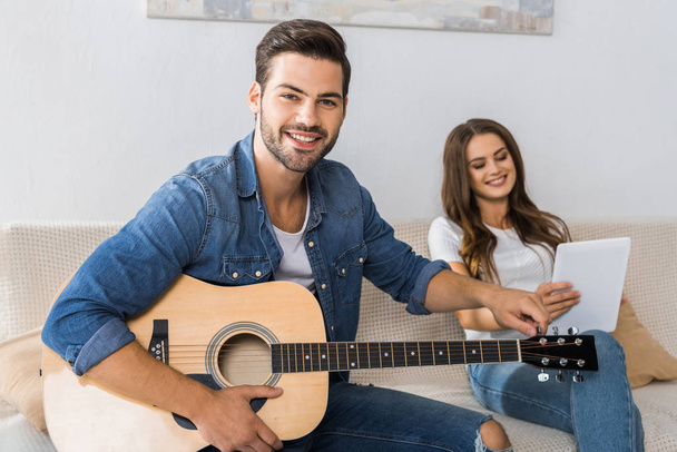 smiling man tuning guitar and looking at camera while his girlfriend sitting near with digital tablet on sofa at home - Foto, Imagem