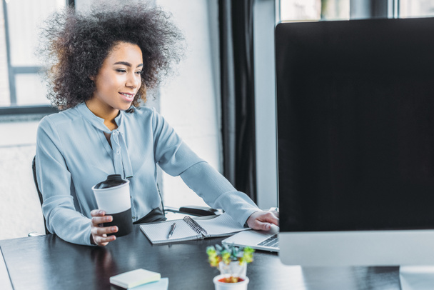 african american businesswoman holding cup of coffee and using laptop in office - Foto, Bild