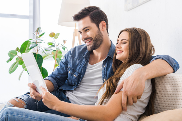 směje se mladý pár sedící na gauči s digitálním tabletu doma  - Fotografie, Obrázek