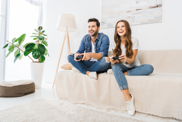 laughing couple playing video game by joysticks on sofa at home  - Foto, immagini