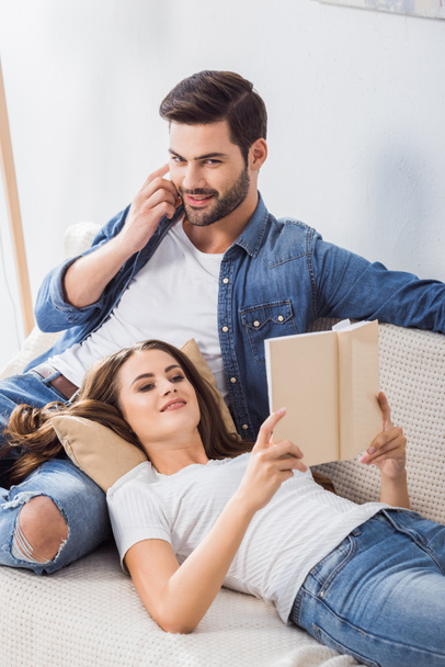 smiling man talking on smartphone while his girlfriend reading book on sofa at home - Photo, Image
