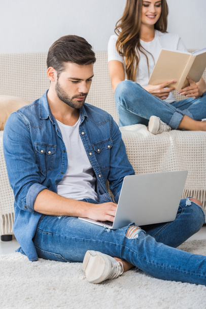 serious man using laptop and sitting on floor while his girlfriend reading book on couch at home - Photo, Image