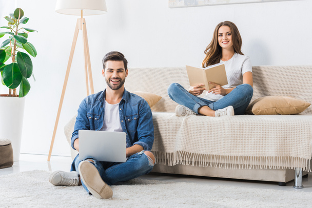 smiling man with laptop sitting on floor while his girlfriend reading book on couch at home - Foto, imagen