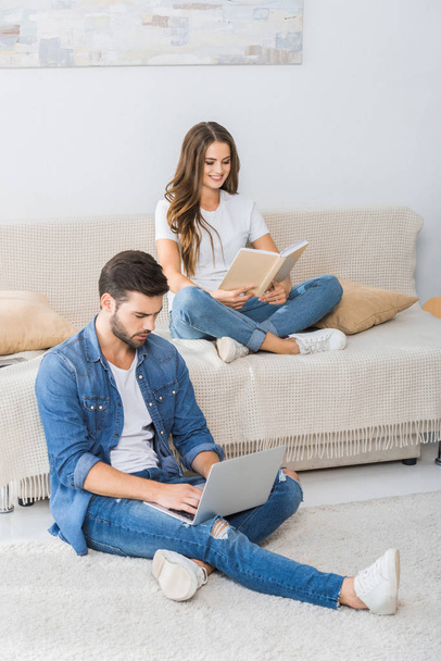 confident man with laptop sitting on floor while his girlfriend reading book on couch at home - Photo, Image
