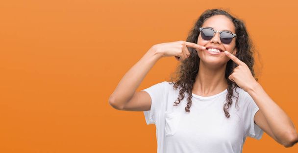 Young hispanic woman wearing sunglasses smiling confident showing and pointing with fingers teeth and mouth. Health concept. - Zdjęcie, obraz