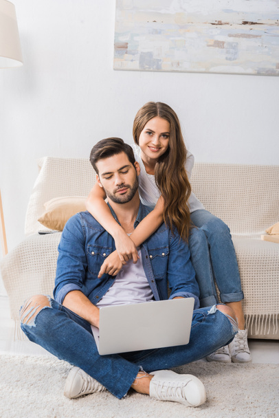 smiling woman embracing boyfriend while he using laptop and sitting on floor at home  - Foto, imagen