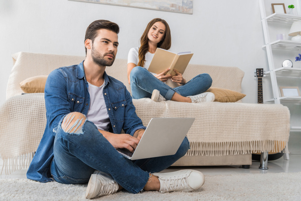 confident young man using laptop and sitting on floor while his girlfriend reading book on couch behind at home - Foto, Imagem