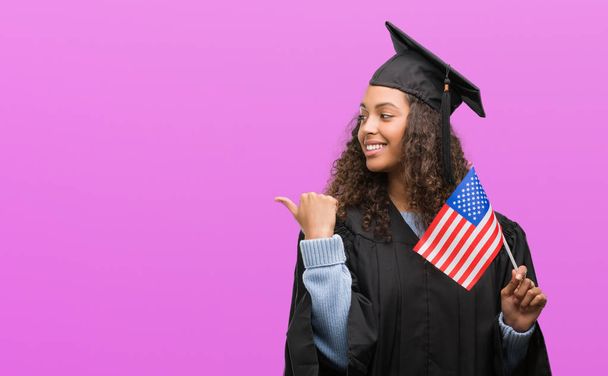 若いヒスパニックの女性を卒業のアメリカ合衆国の国旗を保持している制服を着てポイントと幸せそうな顔を笑顔で側まで親指で示す - 写真・画像