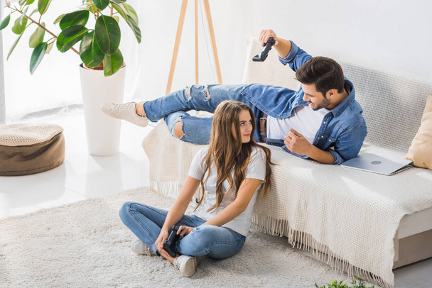 young man threatening by joystick to smiling girlfriend sitting on floor at home   - Photo, image