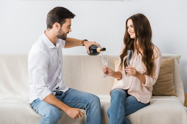 side view of smiling man pouring champagne into glass while sitting on couch with girlfriend at home  - Foto, Imagem