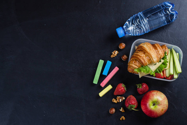 Zurück zum Schulkonzept. Lunchbox mit Croissant, Obst und Gemüse auf schwarzem Hintergrund. Kopierraum, Ansicht von oben - Foto, Bild