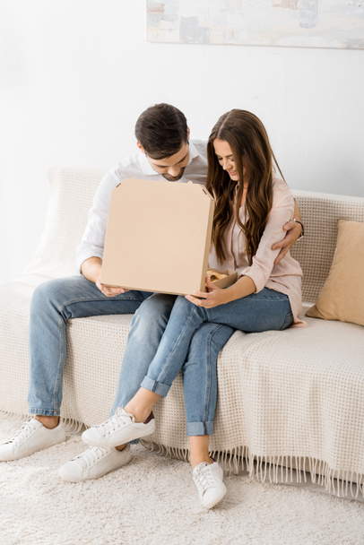 smiling couple with pizza in paper box sitting on sofa at home - Fotografie, Obrázek