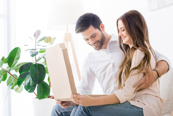 smiling couple with pizza in paper box sitting on sofa at home - Foto, Imagem