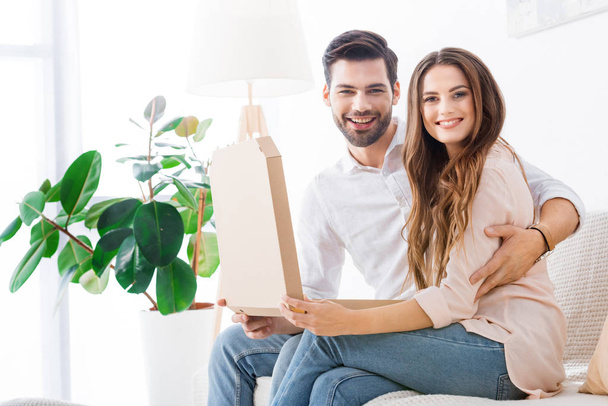 pareja sonriente con pizza en caja de papel sentada en el sofá en casa
 - Foto, Imagen