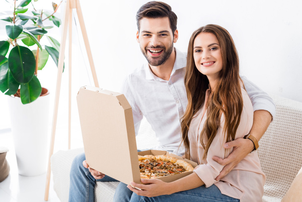 portrait of smiling couple with pizza in paper box sitting on sofa at home - Fotografie, Obrázek