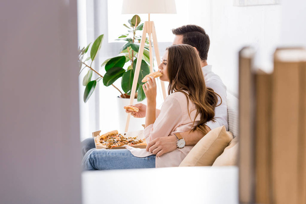 вид збоку молода пара їсть піцу на дивані вдома
 - Фото, зображення