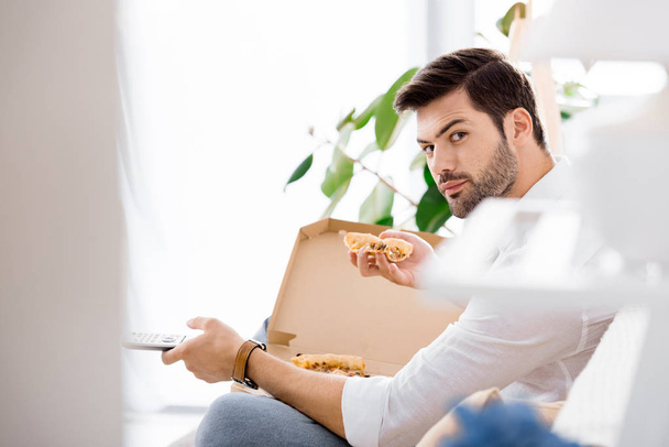 side view of young man with remote control in hand eating pizza alone at home - Photo, image
