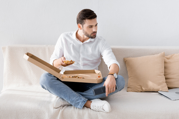 Nuori mies kaukosäätimellä kädessä syö pizzaa yksin kotona
 - Valokuva, kuva