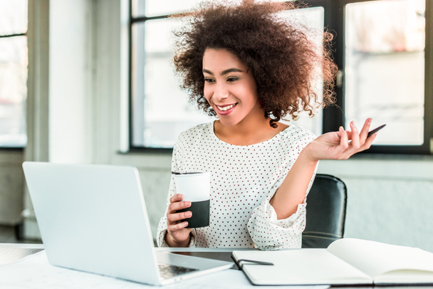 szczęśliwy african american kobieta trzymając kubek kawy i patrząc na laptopie w biurze - Zdjęcie, obraz