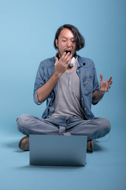 Young bearded man sit down on floor using computer in blue background. Shocked Asian young hipster using laptop, full body shot. Young generation hipster working concept. - Valokuva, kuva
