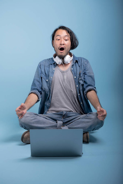 Young bearded man sit down on floor using computer in blue background. Shocked Asian young hipster using laptop, full body shot. Young generation hipster working concept. - Foto, Imagen