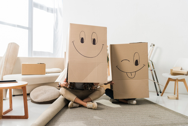 vista oscurecida de pareja con cajas de cartón sobre cabezas sentadas en el piso en casa nueva, concepto de casa móvil
 - Foto, Imagen