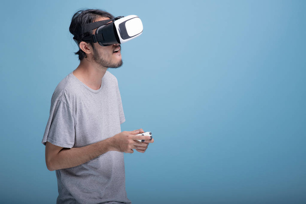Young bearded man playing VR game in blue background. Upset Asian young hipster wearing VR headset, half body shot. Young generation hipster relaxation concept. - Foto, immagini