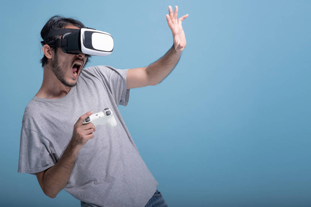 Young bearded man playing VR game in blue background. Extremely scared Asian young hipster wearing VR headset, half body shot. Young generation hipster relaxation concept. - Фото, зображення