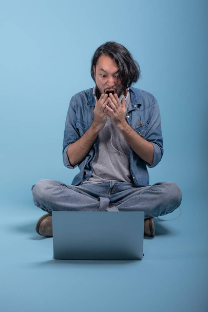 Young bearded man sit down on floor using computer in blue background. Shocked Asian young hipster using laptop, full body shot. Young generation hipster working concept. - Photo, image