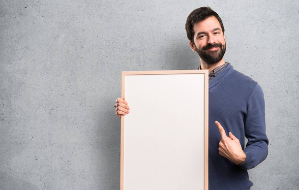 glücklich schöner brünetter Mann mit Bart hält ein leeres Plakat auf strukturiertem Hintergrund - Foto, Bild