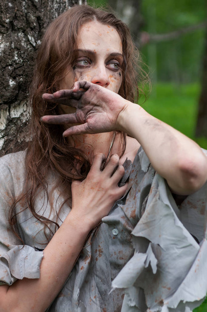 Beautiful young dirty mad and manic looking girl wearing torn clothes and smeared with mud and dried blood stands and huddles to a tree and wipes her face with her hand in the forest. Concept design. - Foto, Imagen