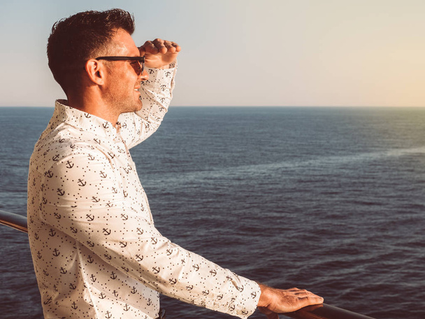 Attractive man in a white shirt with patterns in the form of anchors on the top deck of a cruise ship, looking into the distance on a sunset background. Nautical theme. Concept of sea cruises and rest - Photo, Image