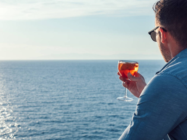 Attractive, stylish man in sunglasses, holding a glass of beautiful pink cocktail on an open deck of a cruise ship on a background of sunset, sea waves and clear sky. Concept of sea travel and recreat - Foto, Bild