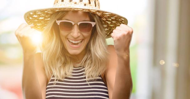 Jeune femme portant des lunettes de soleil chapeau d'été heureux et excité célébrer la victoire exprimant un grand succès, la puissance, l'énergie et les émotions positives. Célébre nouveau travail joyeux
 - Photo, image