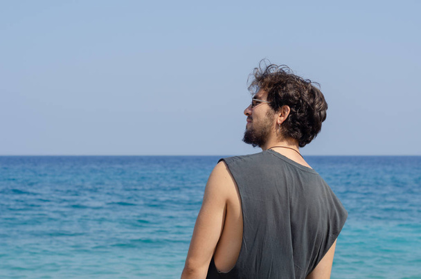 Γενειοφόρος νεαρό άνδρα που φοράει σκουλαρίκι χαμογελά μπροστά στη θάλασσα, ενώ αριστερά του και ψάχνει προς τα εμπρός. - Φωτογραφία, εικόνα