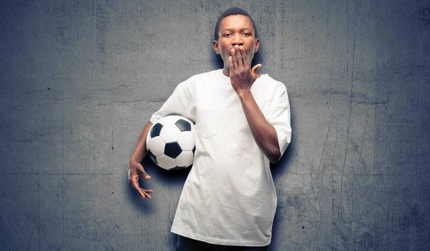 Africký černý muž drží fotbalový míč pokrývá ústa v šoku, vypadá plachý, vyjadřující konceptů mlčení a chybu, strach - Fotografie, Obrázek