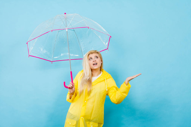 Blonde woman wearing yellow raincoat holding transparent umbrella checking weather if it is raining. - Foto, Imagen