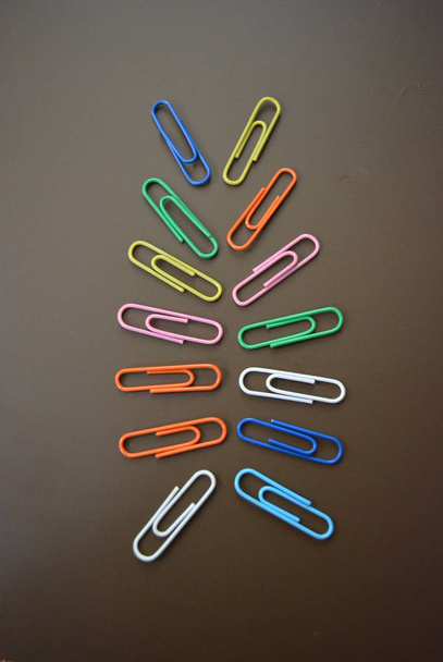 Kleurclips van verschillende kleuren zijn verspreid en veelkleurige clips zijn verspreid over een bruine matte achtergrond. - Foto, afbeelding