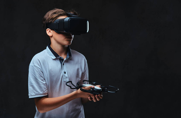 Young boy dressed in a white t-shirt using virtual reality glasses and holds the quadcopter. Isolated on a dark background. - Fotó, kép