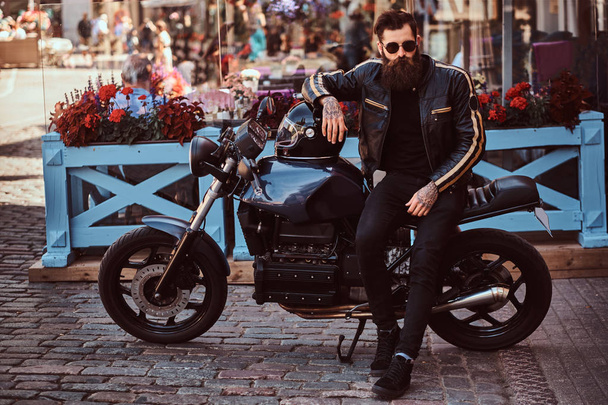 Stylish fashionable biker in sunglasses dressed in a black leather jacket, sitting on his custom-made retro motorcycle near terrace of a cafe. - Photo, Image