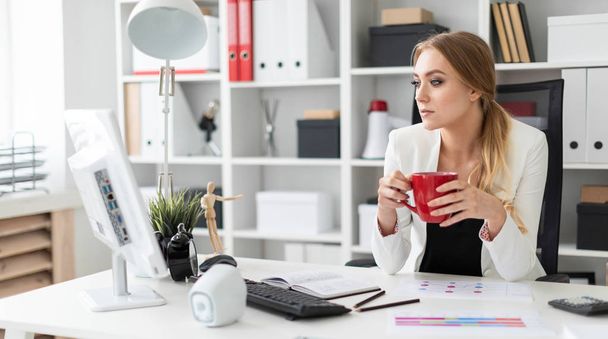 Une jeune fille est assise à un bureau informatique dans le bureau et tient une tasse rouge dans sa main. Avant que la fille sur la table sont des diagrammes
. - Photo, image