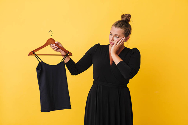 Jonge vrouw in zwarte jurk zorgvuldig bedrijf hanger met zwarte top over gele achtergrond. Plus size model  - Foto, afbeelding