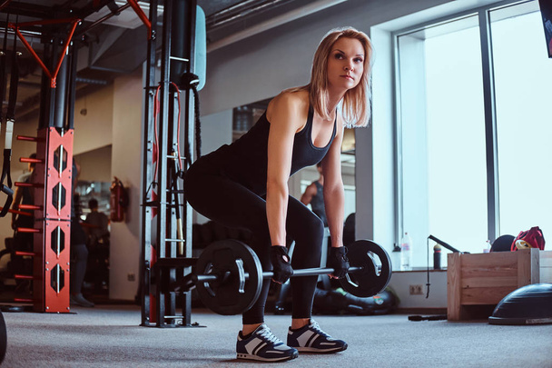 Blonde sportive female in sportswear doing deadlift with barbell in a fitness club or gym. - Photo, Image