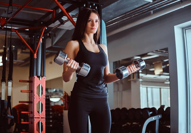 Mulher loira esportiva em sportswear fazendo exercício em bíceps com halteres no clube de fitness ou ginásio
. - Foto, Imagem