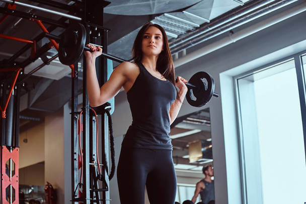 Mooie brunette vrouw in sportkleding houdt een barbell tijdens een training in het fitnesscentrum of sportschool. - Foto, afbeelding