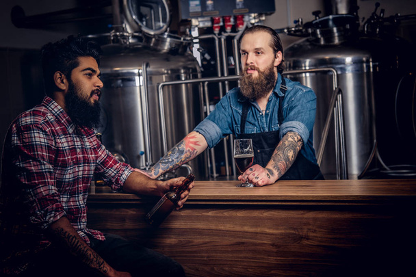 Dwóch przyjaciół Międzyrasowe Brodaty pić piwo w browarze. Hipster Wytatuowany mężczyzna robotnik w fartuch pije piwo z przyjacielem w fabryce browar. - Zdjęcie, obraz